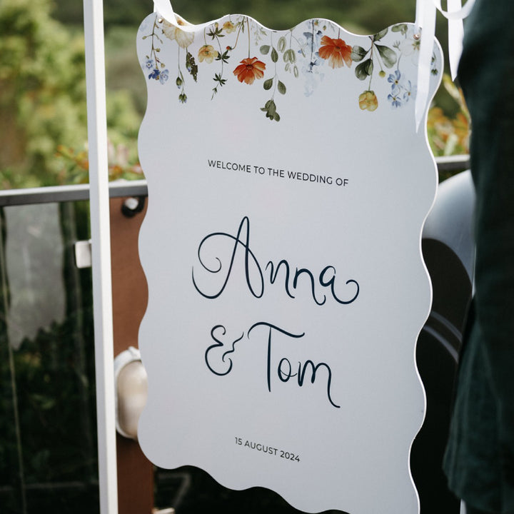 Wavy shape wedding welcome sign with wildflowers and handwritten script fonts.