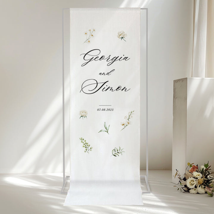 Beautiful calligraphy wedding welcome sign printed on white linen for hanging with white flowers and greenery.