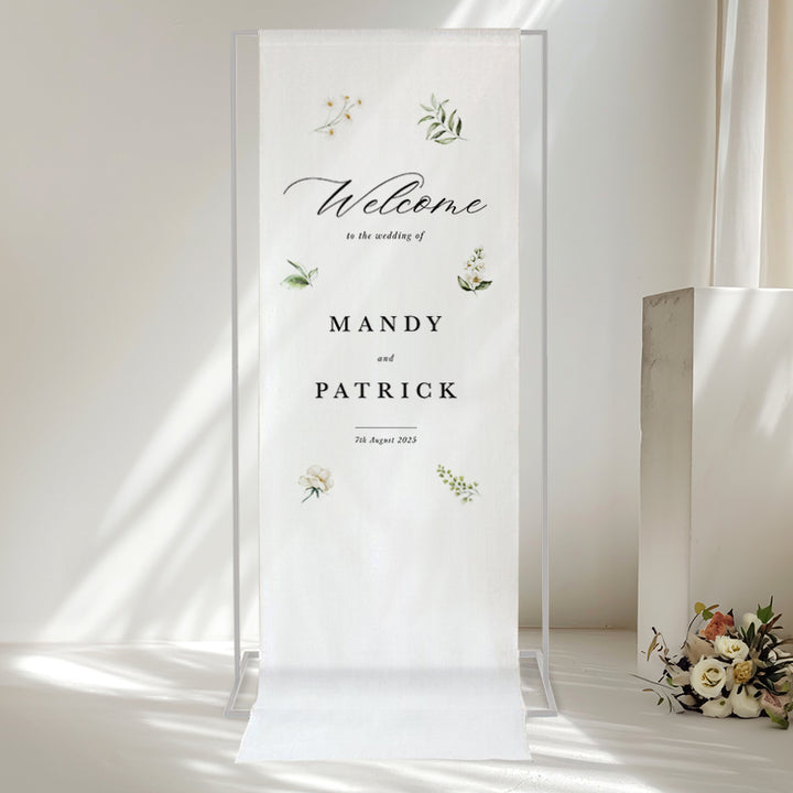 Beautiful calligraphy wedding welcome sign printed on white linen for hanging with white flowers and greenery.